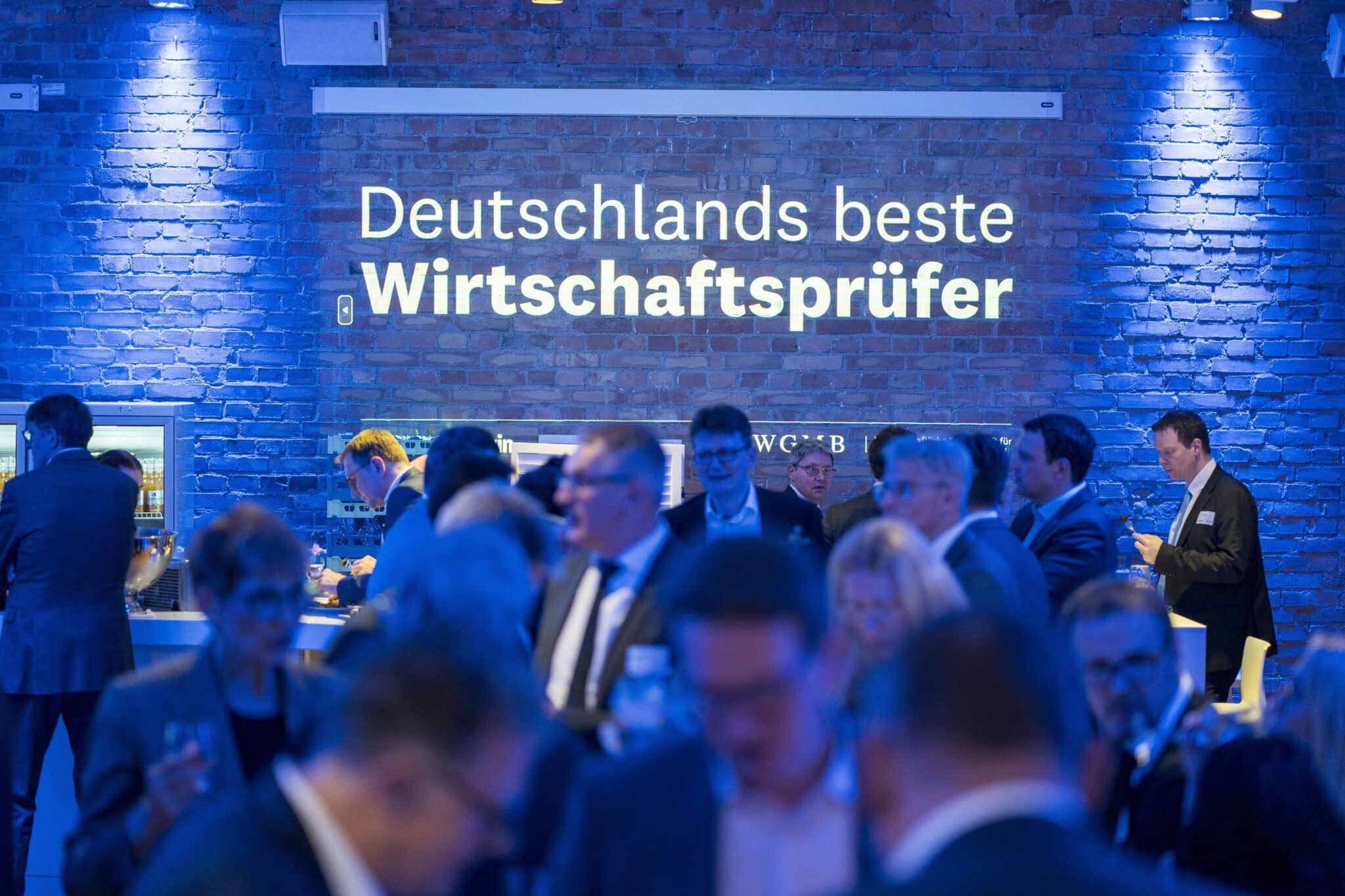Menschenmenge bei einer Veranstaltung mit dem Schriftzug 'Deutschlands beste Wirtschaftsprüfer' an der Wand.