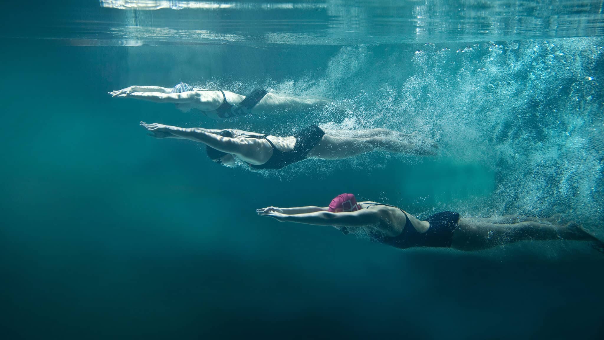 Drei Schwimmerinnen, die synchron unter Wasser tauchen, mit verschiedenen Haarfarben und Schwimmanzügen.