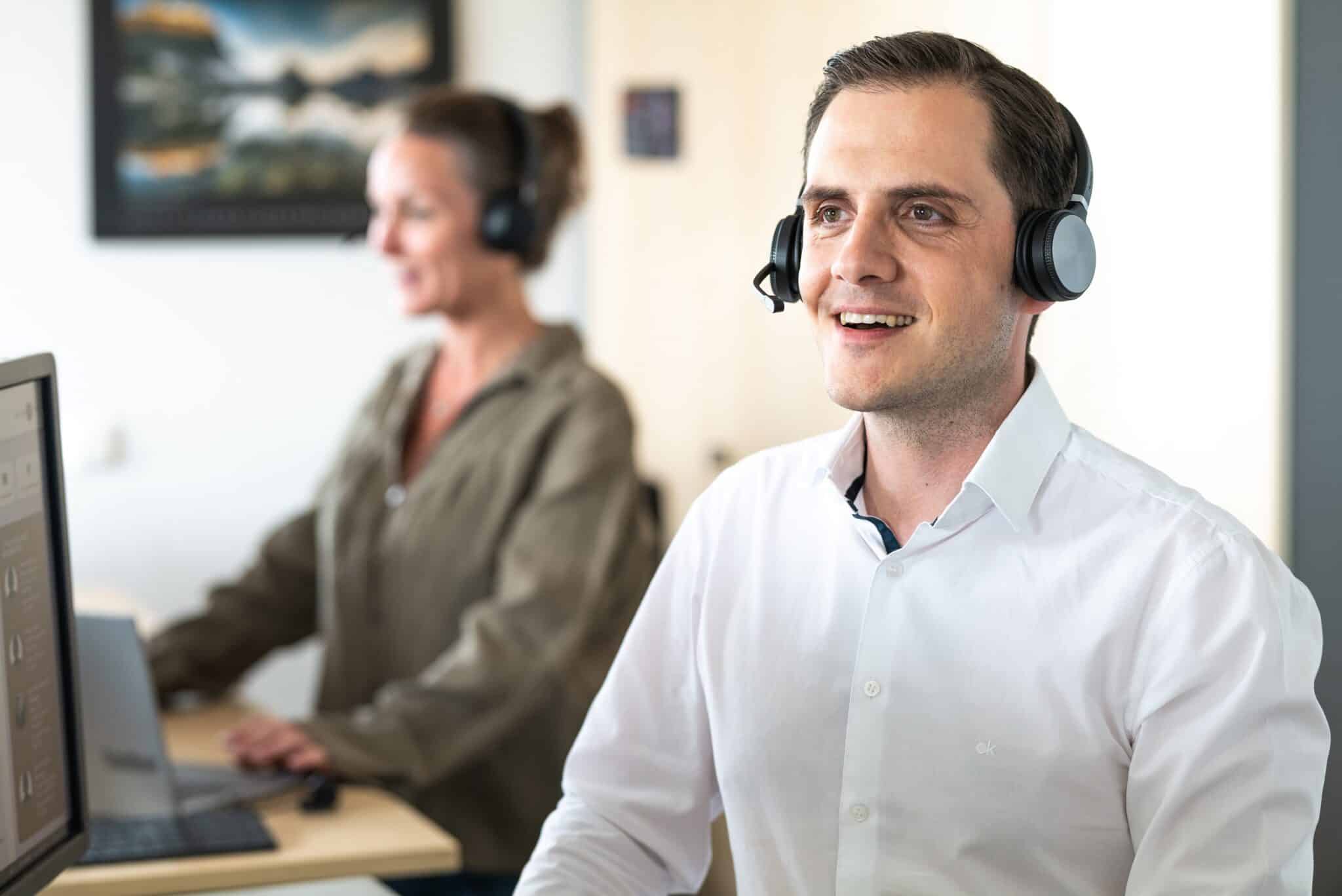 Mann mit Kopfhörern, der an einem Computer arbeitet, während eine Frau im Hintergrund steht.