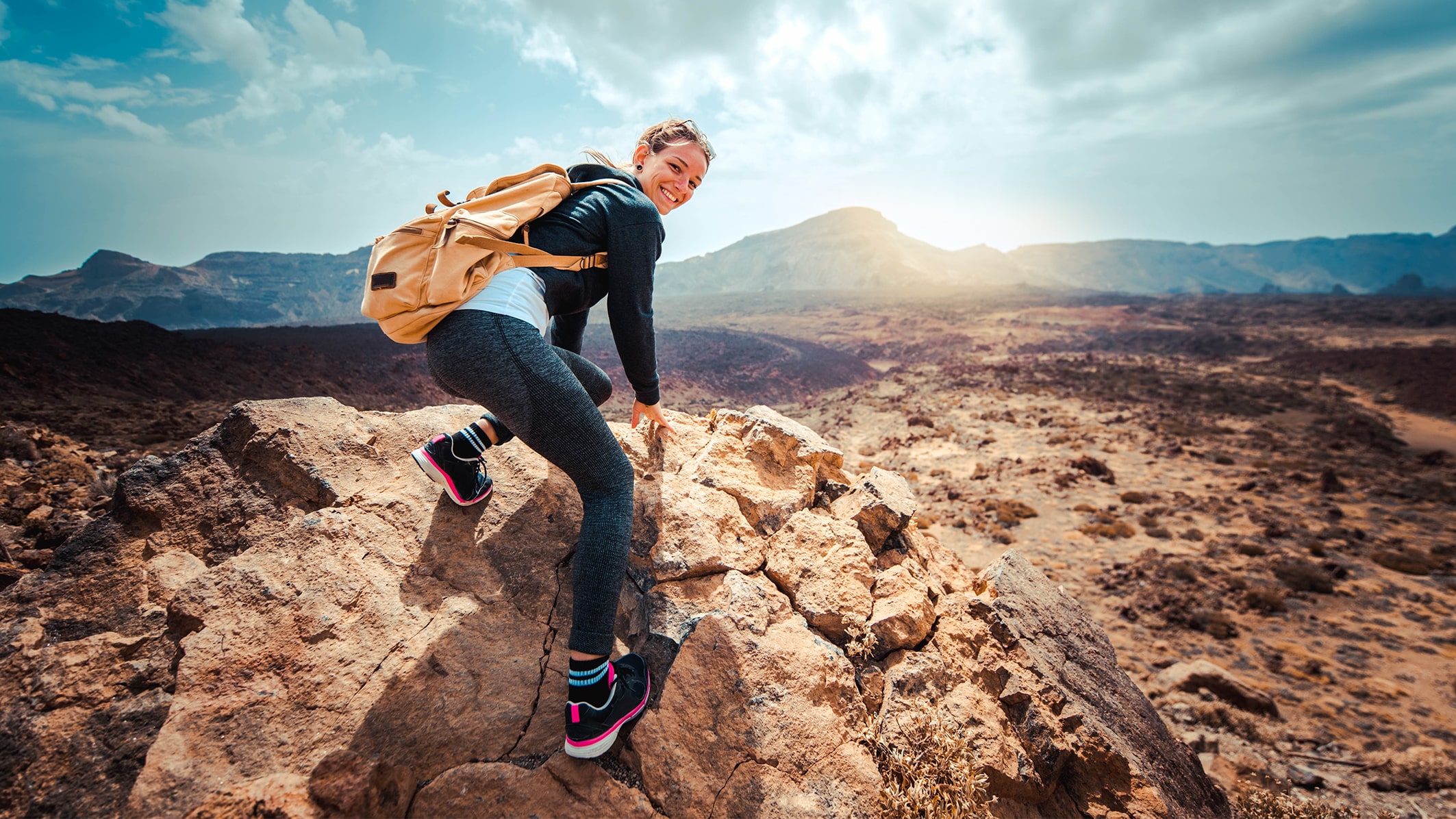 Eine Frau mit einem Rucksack klettert auf einen Felsen in einer bergigen Landschaft.