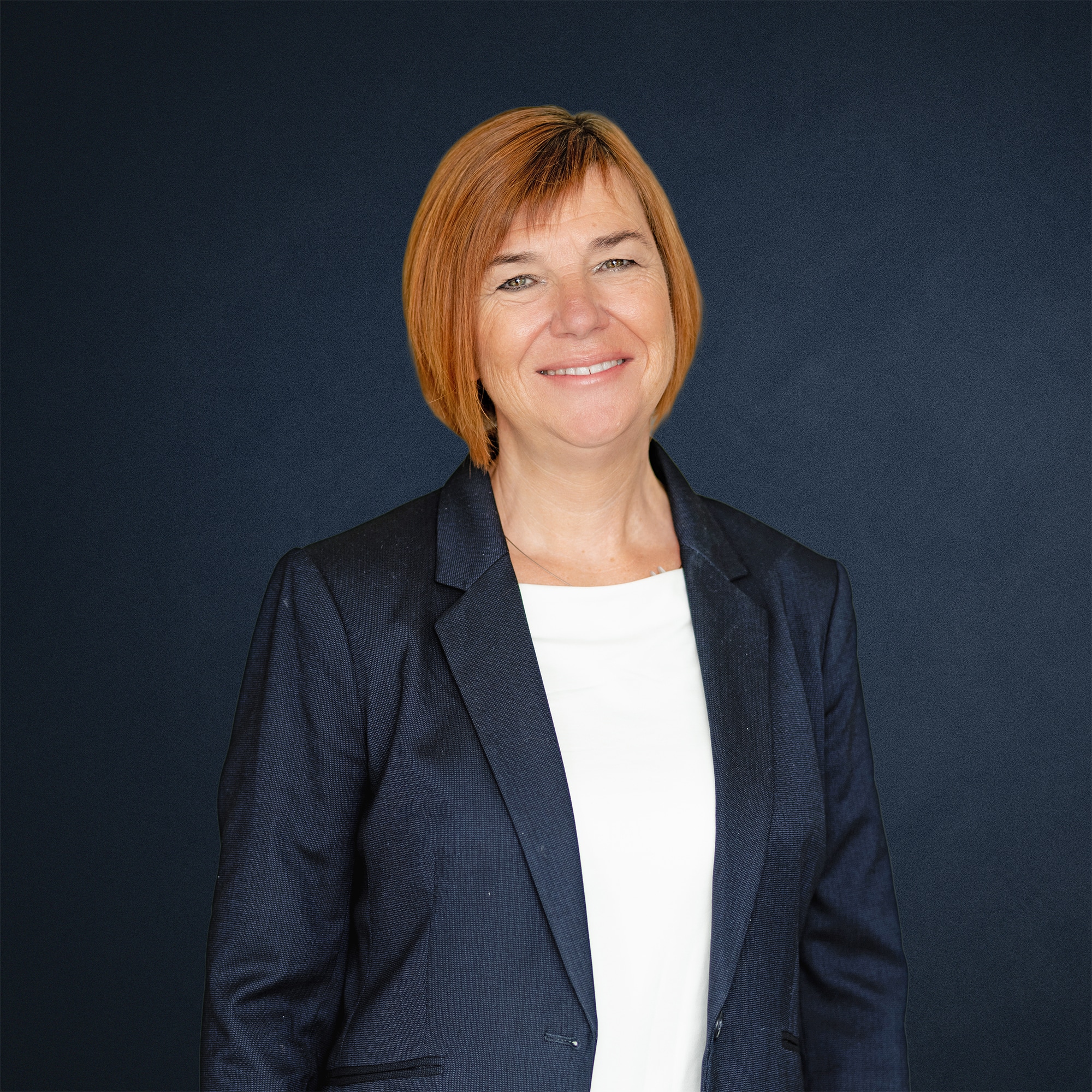 Karin Freischem mit orangefarbenem Haar, trägt einen dunkelblauen Anzug und lächelt vor einem dunklen Hintergrund.