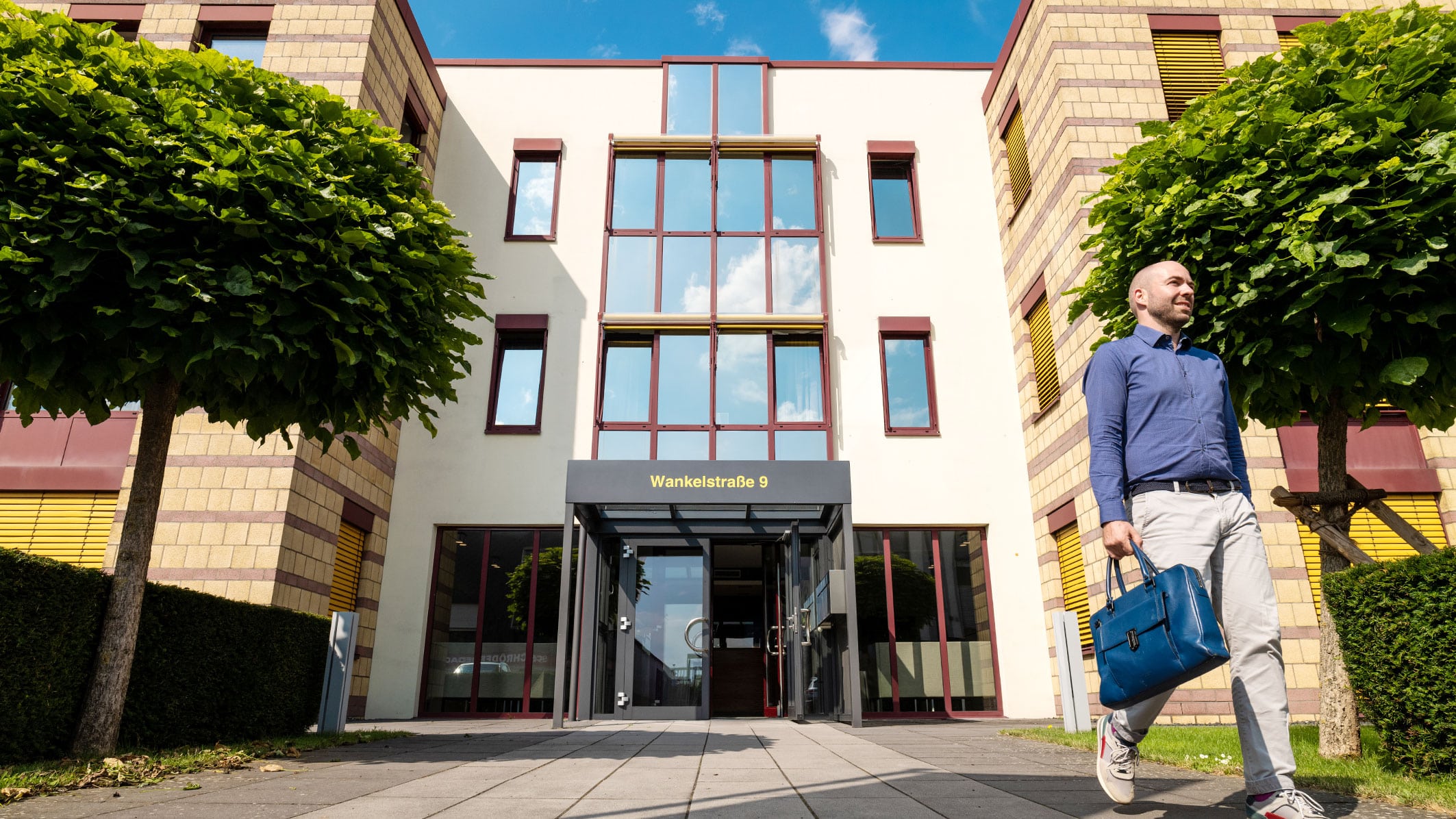 Eingang zu einem modernen Gebäude an der Wankelstraße 9 mit einer Person, die eine blaue Tasche trägt.