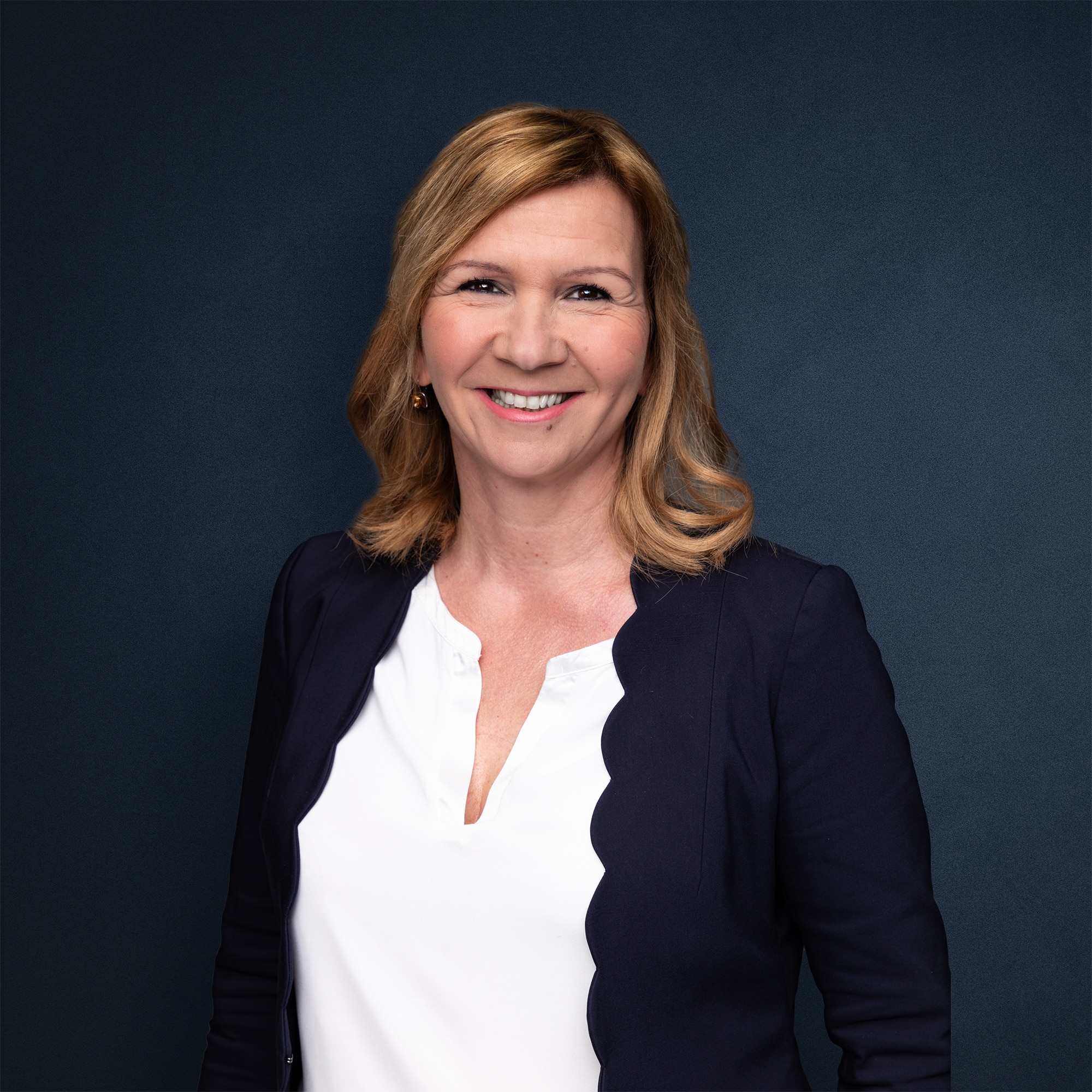 Porträt einer Frau mit blonden Haaren, die in einem blauen Blazer und einem weißen Oberteil lächelt.