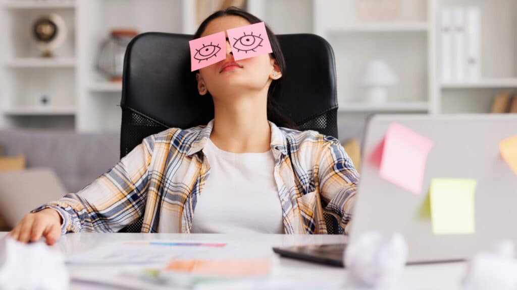Frau sitzt an einem Schreibtisch mit Post-it-Zetteln auf den Augen, umgeben von Notizen und einem Laptop.