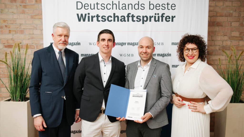 AHW Gruppe präsentiert vier Personen bei der Auszeichnung Deutschlands beste Wirtschaftsprüfer; drei Männer in dunklen Anzügen und eine Frau in weißem Kleid stehen vor Banner, das Manager Magazin thematisiert.