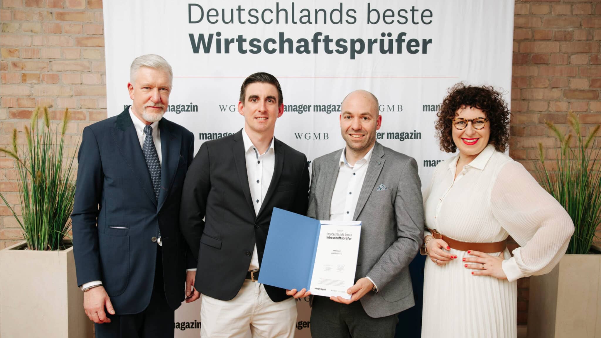 AHW Gruppe präsentiert vier Personen bei der Auszeichnung Deutschlands beste Wirtschaftsprüfer; drei Männer in dunklen Anzügen und eine Frau in weißem Kleid stehen vor Banner, das Manager Magazin thematisiert.